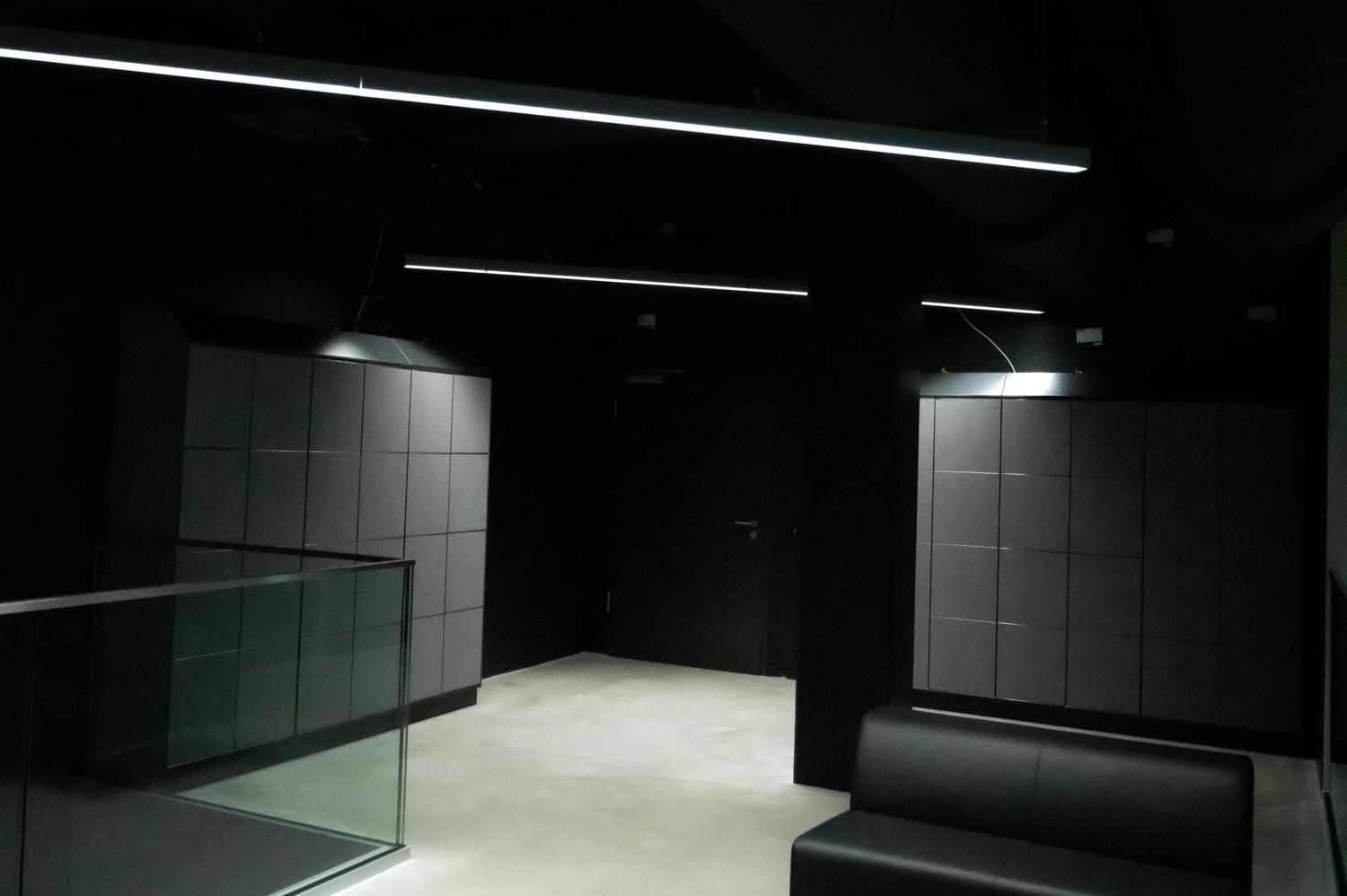 Zwei graue Schließfachblöcke in einem dunklen Foyer mit Glasbrüstung, schwarzem Sofa und linearen LED-Lichtleisten an der Decke