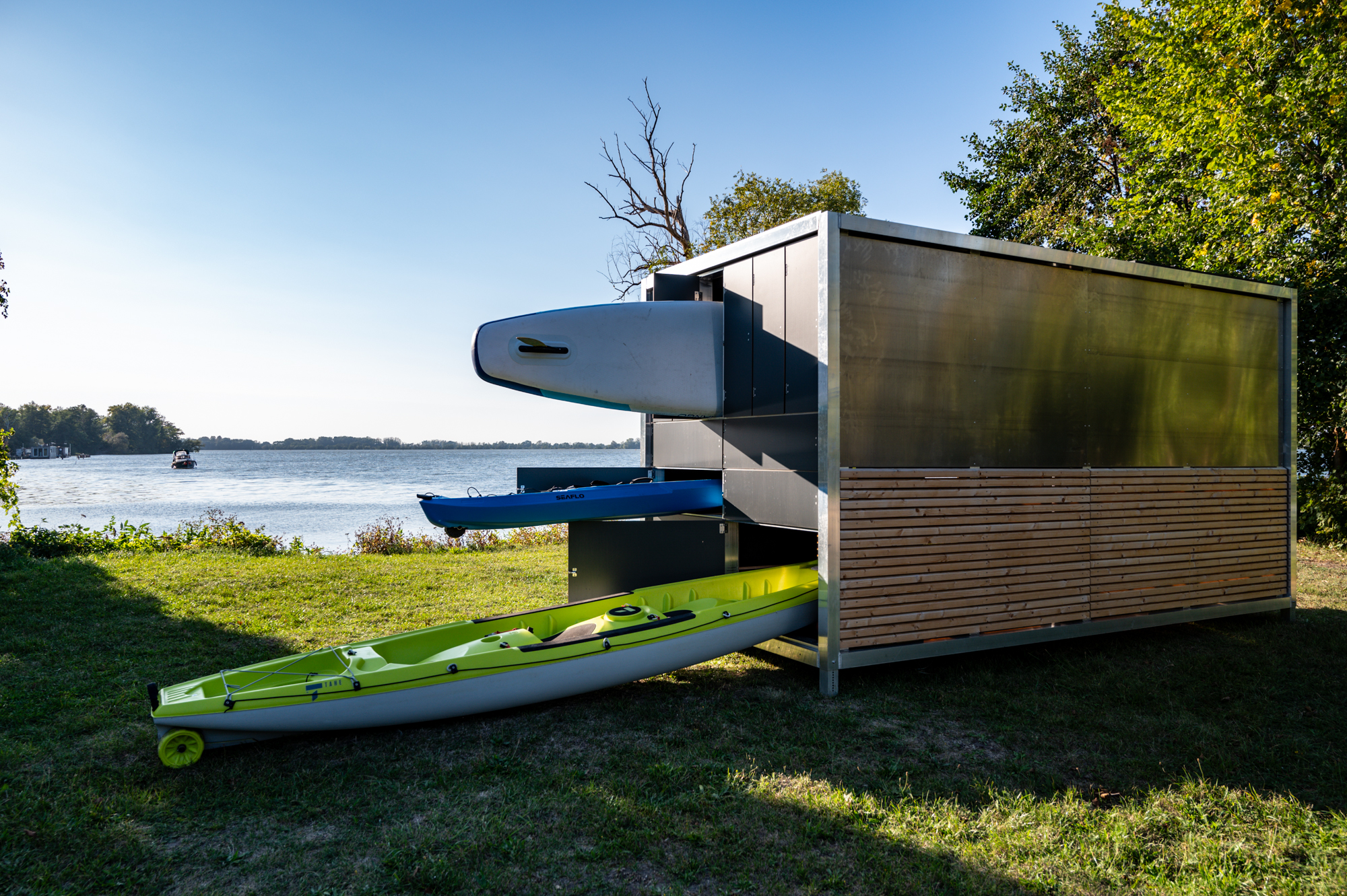 Container mit drei Auszügen am Seeufer: ein Stand-up-Paddleboard und zwei Kajaks, die aus grauen Metallfächern auf eine Wiese geschoben sind